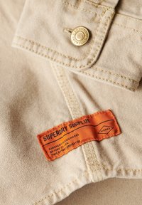 Tela de color beige con una textura suave que cuenta con un botón de presión dorado y una etiqueta naranja que dice "Superdry Surplus" con texto en negro.