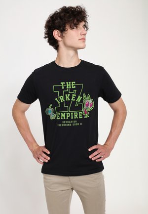 Jonge man met krullend haar, draagt een zwarte T-shirt met groene cartoonfiguren en tekst, handen in de zij, kijkt opzij.