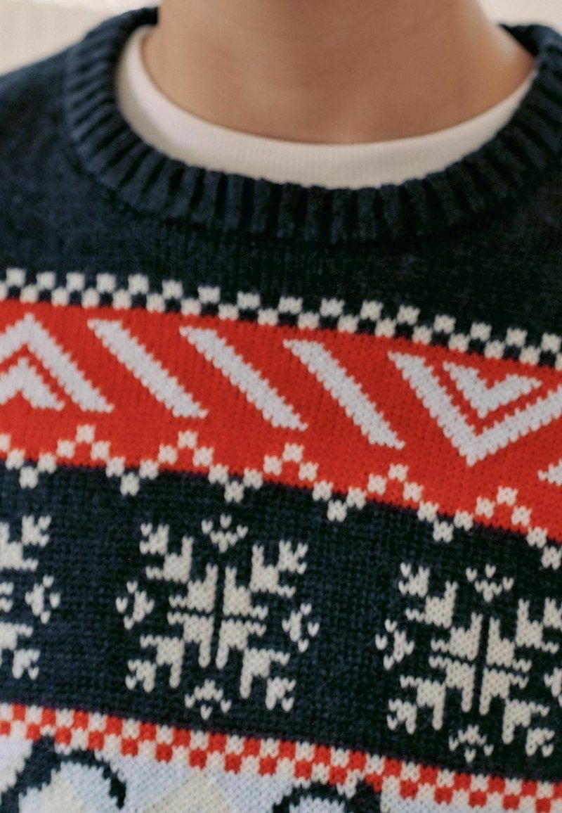 Gros plan d’un pull en laine foncé avec des motifs rouges, blancs et noirs incluant des flocons de neige et des formes géométriques, porté sur une chemise blanche.