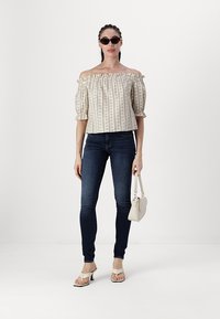 Beige off-the-shoulder-topp med blommönster och puffärmar, kombinerat med mörkblå tajta jeans och vita klacksandaler, hållande en ljus handväska.