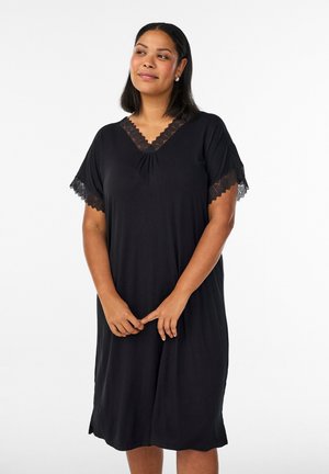 Frau mit mittel-langen Haaren, die ein schwarzes knielanges Kleid mit Spitzenbesatz an Ärmeln und Ausschnitt trägt und mit verschränkten Händen steht.