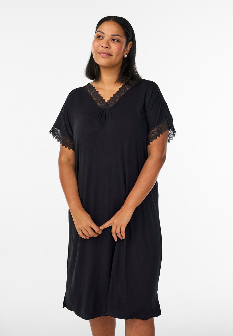 Frau mit mittel-langen Haaren, die ein schwarzes knielanges Kleid mit Spitzenbesatz an Ärmeln und Ausschnitt trägt und mit verschränkten Händen steht.