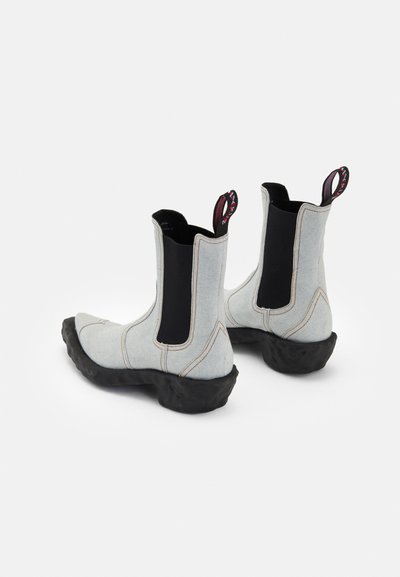 Botines de tobillo de color azul claro hechos de mezclilla con una suela de goma negra. Presentan paneles laterales elásticos y lengüetas de tirón con un diseño único.