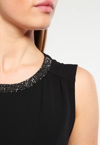Top senza maniche nero con scollatura decorata da perline nere e argento; tessuto morbido con pieghe sottili sulle spalle.