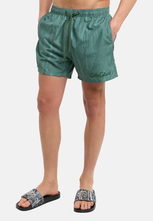 Mand iført grønne shorts med snøre og teksten "Cabo Coluca" samt sorte mønstrede slides, stående med hænderne i lommerne på hvid baggrund.