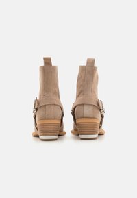 Botines de ante beige con tacón bajo apilado, que presentan correas laterales y acentos de hardware plateado, con un acabado texturizado y suave.