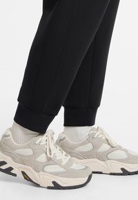 Gray and beige sneakers with mesh and suede materials, featuring a thick sole and padded tongue. Worn with black tapered trousers and white socks.