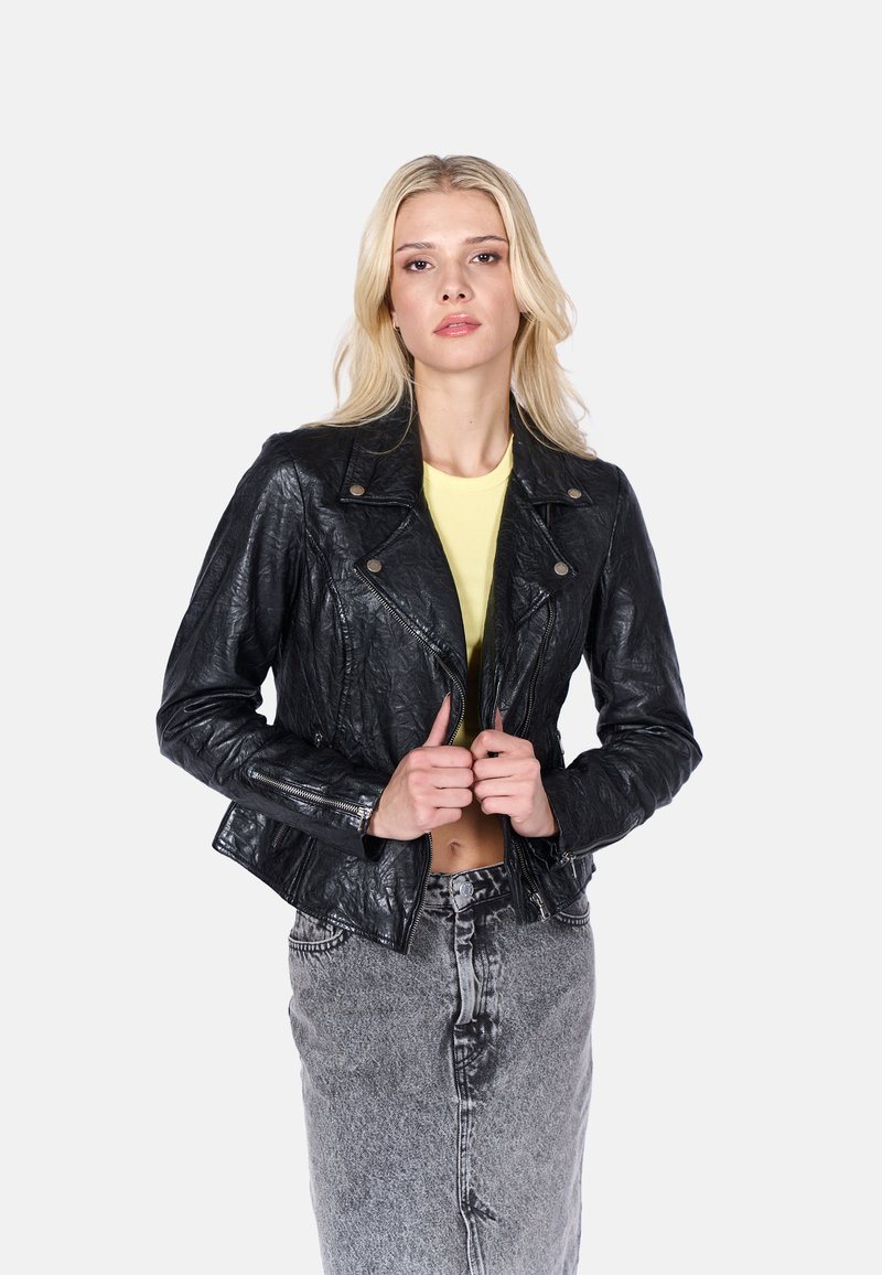 Chaqueta de motociclista de cuero negro, con acabado texturizado, herrajes plateados, bolsillos con cremallera, diseño en capas, combinada con una blusa amarilla y una falda gris.