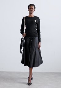 Pulover negru din tricot cu model amplu, accesorizat cu un broșă, asortat cu o fustă A-line neagră cu butoane. Modelul poartă o geantă rotundă neagră și pantofi cu toc negri.