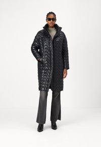Schwarze Pufferjacke mit gewelltem Steppdesign, hohem Kragen und Seitentaschen. Kombiniert mit grauen Schlaghosen und schwarzen Stiefeletten.