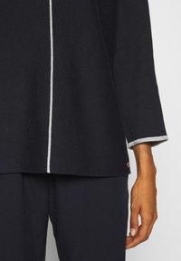 Navyblauer Strickpullover mit weißen vertikalen Nähten, grauen Bündchenakzenten und einem goldenen Logo-Detail am Saum. Der Stoff hat eine strukturierte Optik.