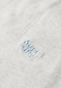 Jasnoszara tkanina o miękkiej teksturze z niebieskim haftowanym logo "Superdry", umieszczonym w widocznym miejscu.