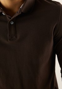 Ruskea poolopaita, jossa on teksturoitu kangas, napillinen halkio ja kontrastivärinen kaulus. Hieman ompeludetaljia kaula-aukon kohdalla.