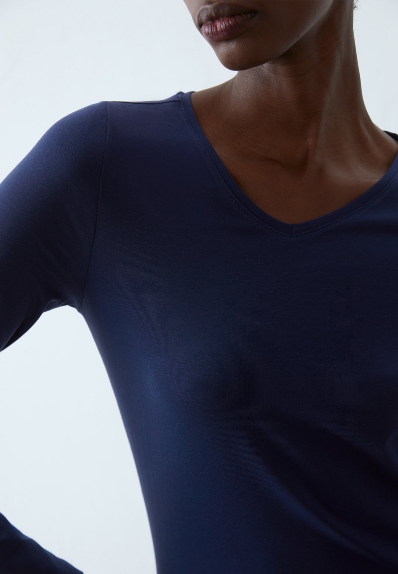 Top blu navy a maniche lunghe con scollatura a V, design aderente, texture liscia e un leggero lucido. I dettagli delle cuciture mettono in risalto le spalle.