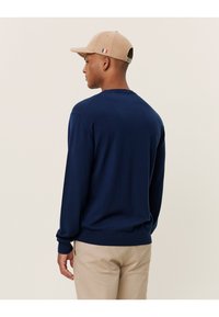 Navyblauer Pullover mit langen Ärmeln, gerippten Bündchen und Saum, von hinten gezeigt. Begleitet von einer beigen Kappe mit einem dreifarbigen Akzent.