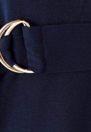 Cinturón de tela azul marino con una gran hebilla circular dorada y textura suave. Cinturón con un acabado cosido y un detalle de diseño en bucle.