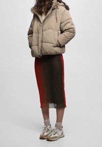 Chaqueta acolchada beige con capucha, sobre una falda midi semitransparente con degradado en tonos marrón, rojo y negro. Calzado ligero con acentos pastel.