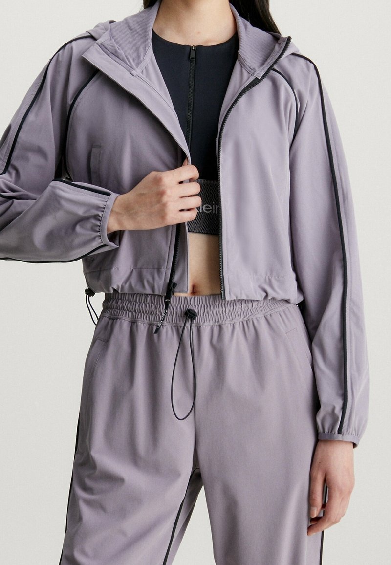 Femme portant un survêtement court lavande avec des bordures noires, en train de dézipper la veste pour révéler un soutien-gorge de sport noir en dessous.