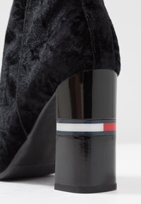 Schwarze Stiefeletten mit einer strukturierten, plüschigen Außenseite und einem glänzenden schwarzen Blockabsatz. Der Absatz hat ein Band mit rot, weiß und blau gestreiften Mustern.