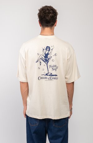 Camiseta de algodón color crema con un corte relajado. El gráfico en la parte posterior presenta a una mujer en una copa de martini, el texto dice "¡Salud por la Noche del Viernes, Queridos!"