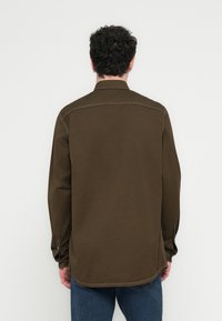 Man som bär en mörk olivgrön långärmad tröja och blå jeans, stående med ryggen mot kameran mot en enfärgad vit bakgrund.