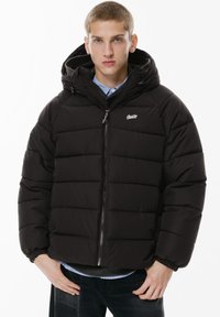 Sort dunjakke med quiltet design, frontlynlås, justerbar hætte og elastiske manchetter, båret over en skjorte med krave og jeans.