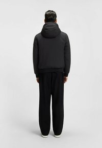 Man står med ryggen mot kameran iförd en svart vadderad jacka med huva, lösa svarta byxor och vita skor framför en enkel grå bakgrund.