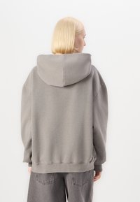 Sweatshirt supradimensionat, de culoare gri, cu o glugă mare, manșete și tiv cu nervuri, realizat dintr-un material moale. Are o croială lejeră și detalii minime.