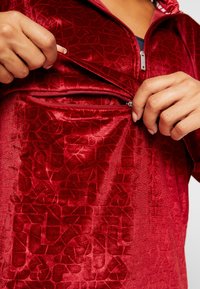 Haut rouge zippé en tissu texturé avec un motif géométrique. Dispose d'une poche ventrale avec une fermeture éclair métallique et d'un col montant.