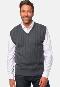 Lächelnder Mann, der einen dunkelgrauen, grob gestrickten, ärmellosen Pullover über einem weißen Hemd mit Knöpfen und dunklen Jeans trägt, vor einem schlichten hellen Hintergrund.