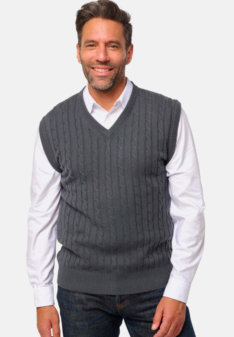 Lächelnder Mann, der einen dunkelgrauen, grob gestrickten, ärmellosen Pullover über einem weißen Hemd mit Knöpfen und dunklen Jeans trägt, vor einem schlichten hellen Hintergrund.