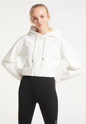 Jonge vrouw met blond haar die een witte cropped hoodie en zwarte high-waisted legging draagt, staat met handen in de zij.