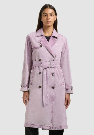 Kobieta ubrana w lawendowy płaszcz trencz z paskiem, zapinany na dwa rzędy guzików, z mankietami na guziki i szerokim kołnierzem, stojąca na jednolitym tle.
