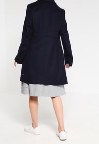 Mörkblå ullblandad kappa med strukturerad krage och långa ärmar, med rak skärning och subtil märkning nära fållen.