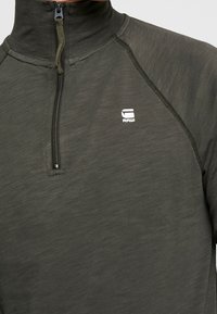 Ciemnozielony polar z zamkiem błyskawicznym i wysokim kołnierzem, wykonany z lekkiej tkaniny. Posiada małe białe logo na piersi oraz płaskie szwy.