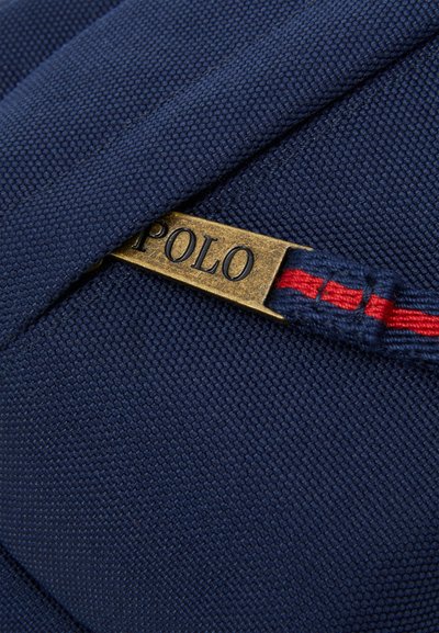 Ναυτικό ύφασμα τσάντας με υφή επιφάνειας, διαθέτει μεταλλικό φερμουάρ με χαραγμένο το "POLO", ενισχυμένο από μια κόκκινη και μπλε ριγέ θηλιά.