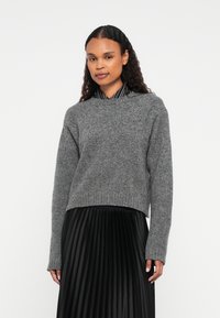 Donna con capelli ricci che indossa un maglione grigio a maglia sopra una gonna plissettata nera e una camicia a righe bianco e nere con colletto.