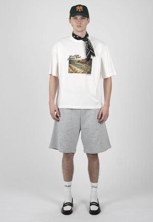Junger Mann mit weißem T-Shirt mit Grafik, grauen knielangen Shorts, schwarz-weißen Loafers, weißen Socken, schwarzem Bandana und dunkelgrünem Cap.