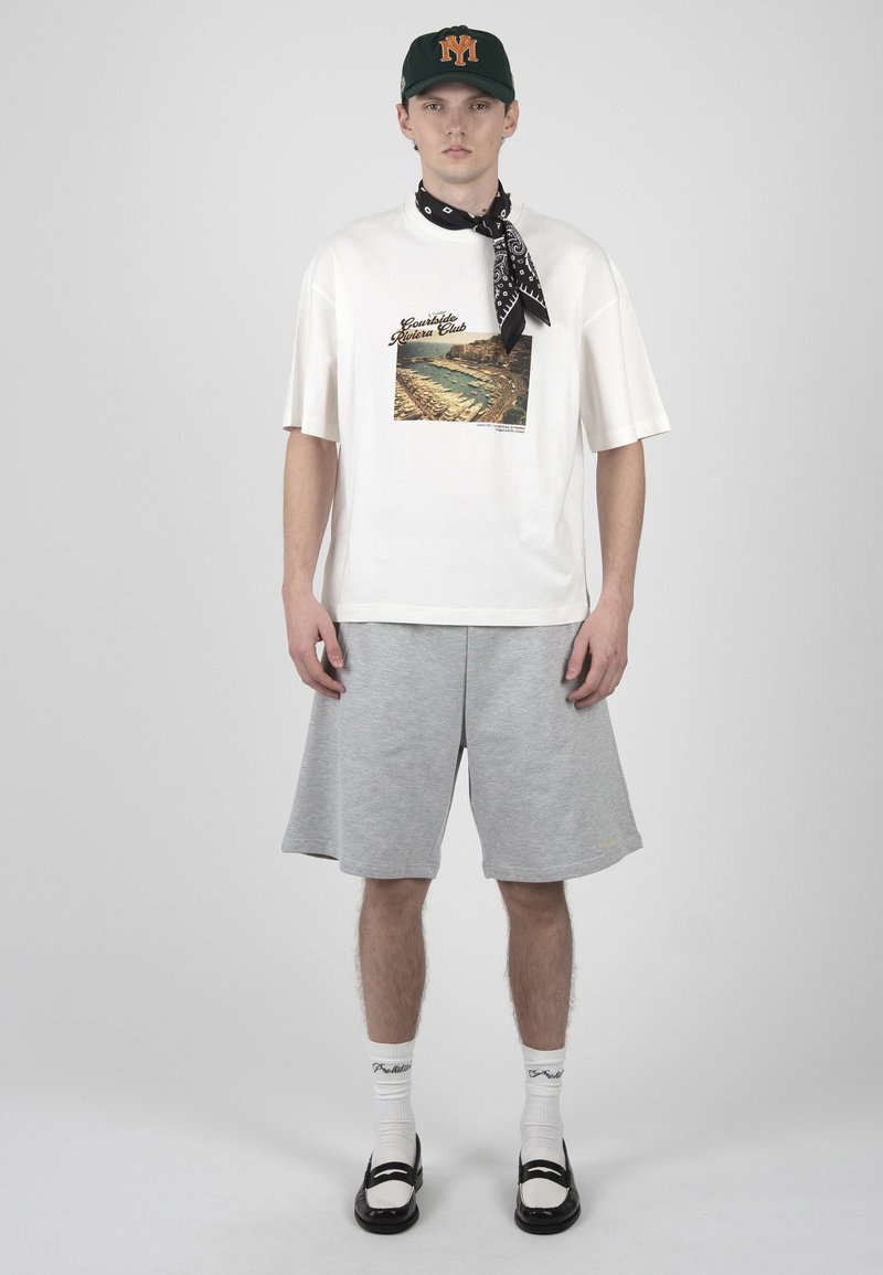 Jeune homme portant un t-shirt blanc à motifs, un short gris mi-cuisses, des mocassins noirs et blancs, des chaussettes blanches, un bandana noir et une casquette vert foncé.
