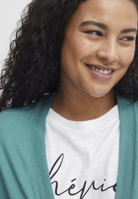 En turkos kofta med ribbat mönster som bärs över en vit skjorta med svart kursiv text. Mjukt tyg med avslappnad passform.