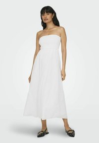 Vestido blanco sin mangas con un cuerpo fruncido, silueta maxi fluida y tela ligera. Usado con zapatillas negras con tachuelas decorativas.