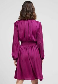 Robe fuchsia à manches longues, taille smockée, jupe fluide et texture douce. L'arrière présente un tissu froncé pour un détail supplémentaire.