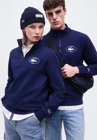 Deux jeunes adultes portant des pulls Lacoste bleu marine à zip quart avec logos, la femme porte également un bonnet assorti, l'homme porte un sac bandoulière noir.
