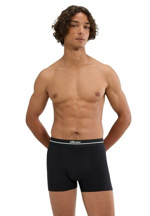 Młody mężczyzna z kręconymi włosami prezentujący czarne bokserki Ellesse, stojący z rękami za plecami na białym tle.
