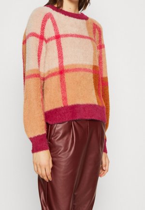 Femme portant un pull à carreaux duveteux en tons beige, rose et orange, associé à un pantalon en cuir bordeaux taille haute, debout devant un fond blanc.