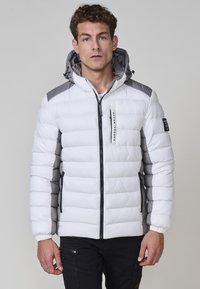 Giacca bianca soffice con accenti grigi, dotata di una chiusura con zip, cappuccio e polsini elasticizzati. Il design include sezioni trapuntate verticali.