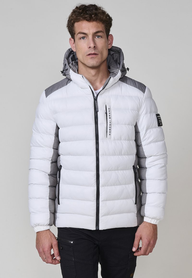 Giacca bianca soffice con accenti grigi, dotata di una chiusura con zip, cappuccio e polsini elasticizzati. Il design include sezioni trapuntate verticali.