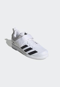 Zapatilla deportiva blanca con parte superior de malla, tres rayas negras y una correa de velcro. Presenta un material texturizado y una suela de goma para agarre.
