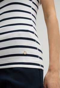 Witte geribde tanktop met marineblauwe horizontale strepen, met een ronde zilveren logo-accent bij de zoom. Gecombineerd met donkerblauwe broek.