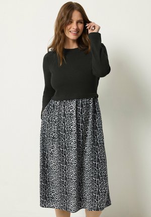 Mujer con cabello castaño que lleva una blusa negra de manga larga y una falda estampada de leopardo en blanco y negro, sonriendo con una mano cerca de su rostro.
